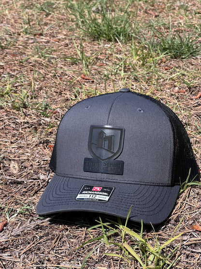 A dark gray and black trucker hat with a black mesh back and a black shield logo on the front, resting on a patch of dry grass and dirt outdoors.