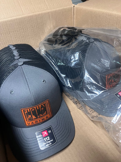 A dark gray and black trucker hat with an orange and black patch on the front, displayed next to another hat in clear plastic packaging inside a cardboard box.