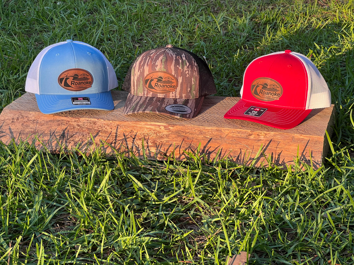Three trucker-style baseball caps displayed outdoors on a wooden board in grass. The left cap is light blue and white, the center cap has a brown camouflage pattern, and the right cap is red and white.