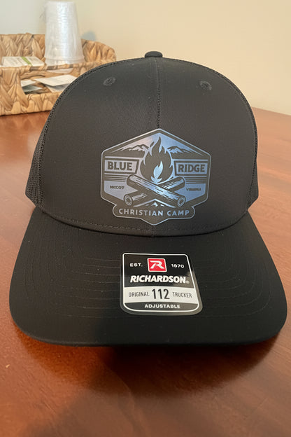 A black Richardson 112 trucker hat with a blue and gray 'Blue Ridge Christian Camp' patch on the front and a Richardson brand sticker on the visor, resting on a wooden table.
