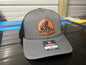 A gray and black Richardson 112 trucker hat with a circular brown leather patch on the front that reads 'QUALITY WELDING & FABRICATION'. The hat is resting on a dark reflective surface with a workshop background.